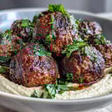 Frying venison meatballs with aromatic spices in a skillet beside fresh salad and hummus.