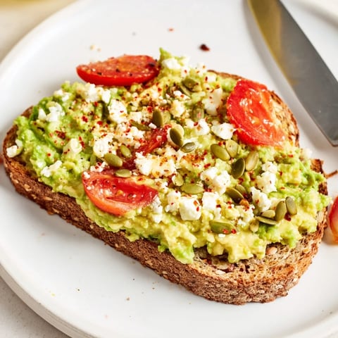 Two slices of avocado toast on whole grain bread topped with fresh tomato and crumbled feta cheese.